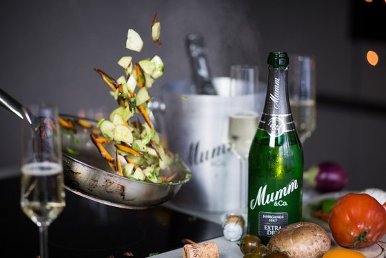 Ein Kochprozess wird festgehalten: Gemüse wird in die Luft geworfen, umgeben von Sekt und frischen Zutaten.