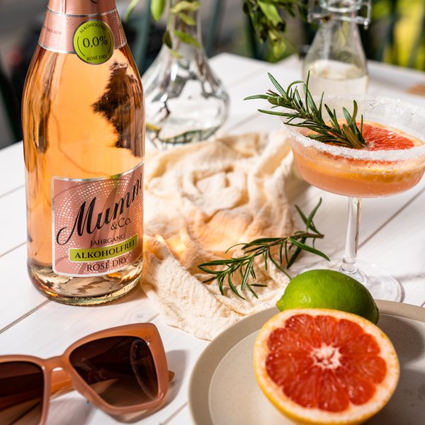 Eine Flasche alkoholfreier Rosé-Sekt steht auf einem Tisch, umgeben von frischen Kräutern und einer stilvollen Cocktailglas.