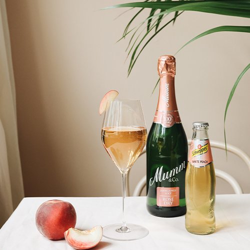 Ein stilvolles Arrangement mit einem Glas Rosé-Sekt, einer Flasche Mumm und einem frischen Pfirsich auf einem weißen Tisch.