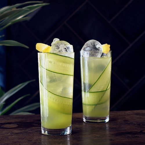 Zwei hohe Gläser mit einem erfrischenden Getränk, gefüllt mit Eis, Gurkenscheiben und Zitronenstücken, vor dunklem Hintergrund.