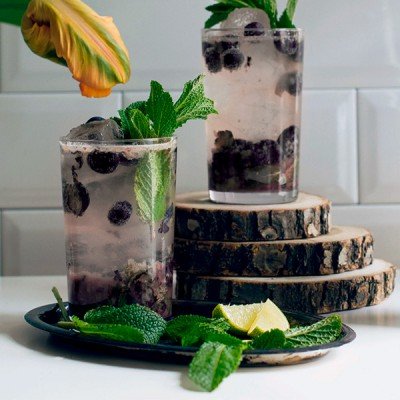 Zwei Gläser mit einem erfrischenden Getränk aus Blaubeeren, Minze und Eiswürfeln stehen auf einem rustikalen Holzuntergrund.