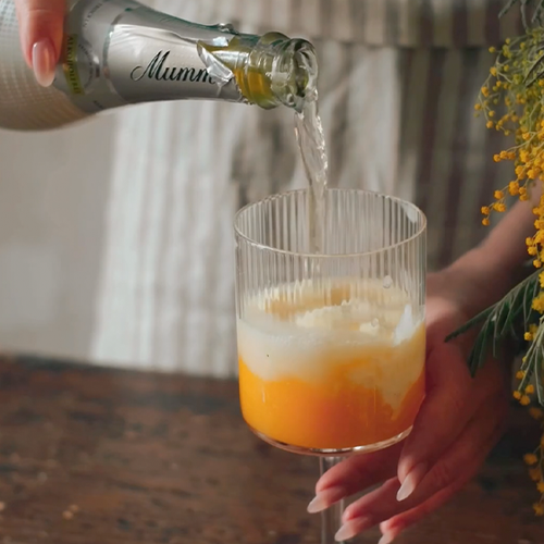 Ein Glas mit orangefarbenem Getränk wird mit sprudelndem Sekt gefüllt, während eine Hand das Glas hält.