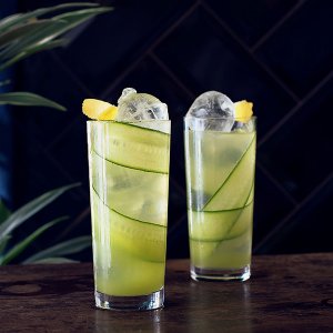 Zwei hohe Gläser mit einem erfrischenden Getränk, gefüllt mit Eis, Gurkenscheiben und Zitronenstücken, vor dunklem Hintergrund.