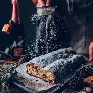 Ein frisch gebackener Stollen wird mit Puderzucker bestäubt, während eine Flasche Sekt im Hintergrund steht.