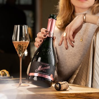 Die Frau mit blonden Haaren zeigt eine Flasche Mumm Sekt und ein Glas mit sprudelndem Rosé, umgeben von einem stilvollen Ambiente.