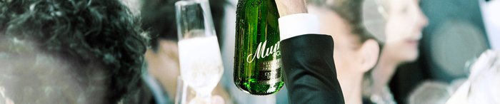 Eine Hand hebt eine grüne Flasche Sekt, während im Hintergrund verschwommene Gesichter von feiernden Menschen zu sehen sind.