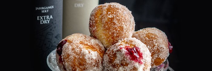 Zuckrige Krapfen in einer Schale, die mit einer dunklen, stilvollen Sektflasche im Hintergrund harmonieren.