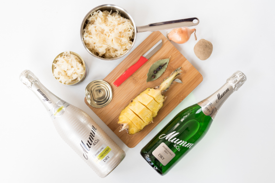 Zwei Flaschen Mumm Sekt liegen neben einer Holzplatte mit frischer Ananas, Zwiebel und Sauerkraut.