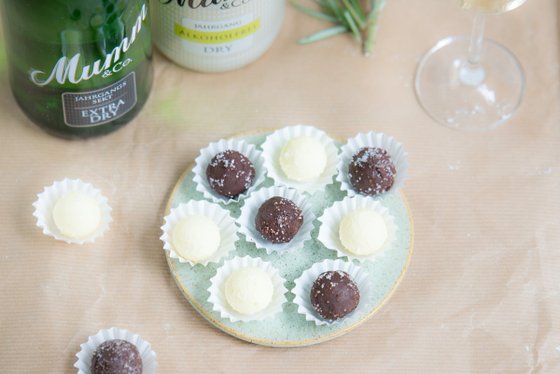 Eine Auswahl an Pralinen in Weiß und Dunkelbraun, dekoriert mit Zucker, auf einem grünen Teller neben Sektflaschen.
