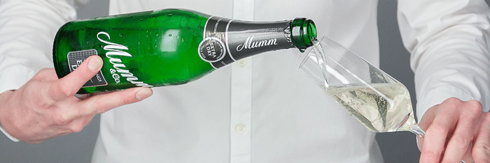 Eine Person in einem weißen Hemd gießt Sekt aus einer grünen Flasche in ein elegantes Glas.