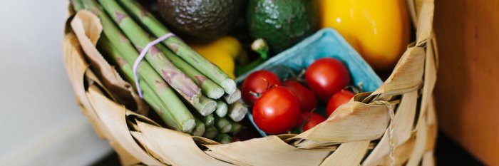 Ein geflochtener Korb voller frischer Gemüse, darunter grüne Spargel, Tomaten und Avocados, strahlt gesunde Farben aus.