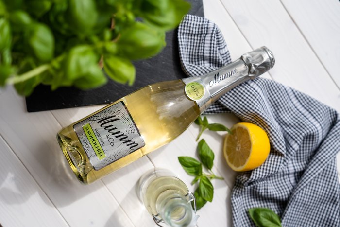 Eine Flasche alkoholfreier Sekt steht auf einem weißen Tisch, umgeben von frischem Basilikum und einer halbierten Zitrone.