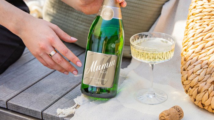 Eine Hand hält eine Flasche Mumm Sekt, während ein Glas mit sprudelndem Getränk auf einem Tisch steht.