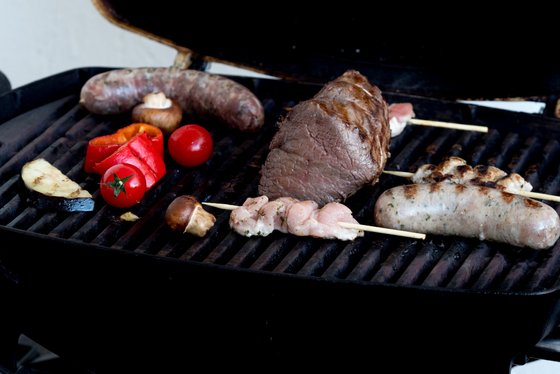 Auf dem Grill liegen verschiedene Fleischsorten, darunter Würste und Spieße, umgeben von buntem Gemüse wie Paprika und Tomaten.