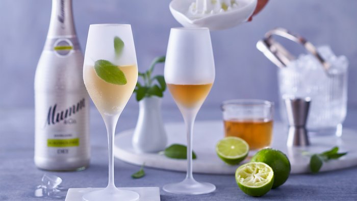 Eine Flasche Mumm-Sekt und zwei Gläser mit einem erfrischenden Cocktail, umgeben von Limetten und Minze, laden zum Genießen ein.