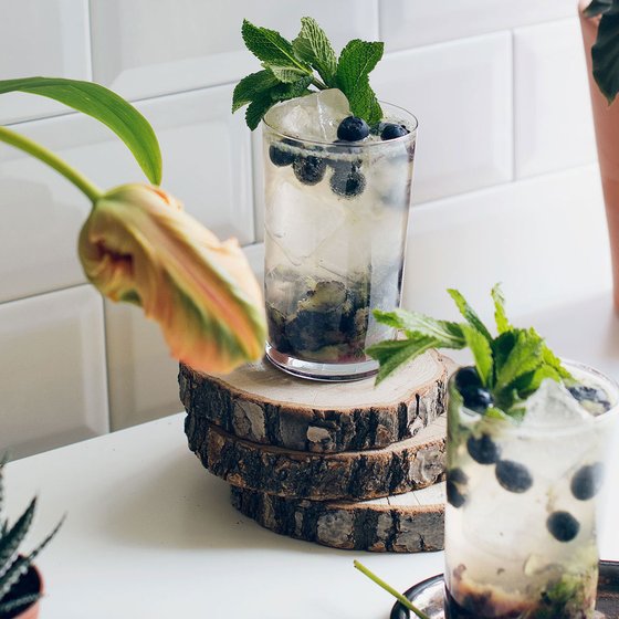 Zwei Gläser mit einem erfrischenden Getränk, gefüllt mit Eis, Minze und Heidelbeeren, stehen auf einem Holzuntersetzer.