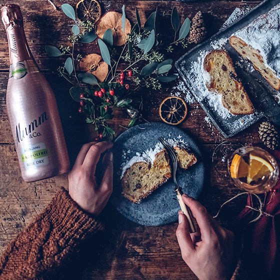 Eine Hand schneidet ein Stück Stollen auf einem blauen Teller, umgeben von festlichen Dekorationen und einem Glas Sekt.