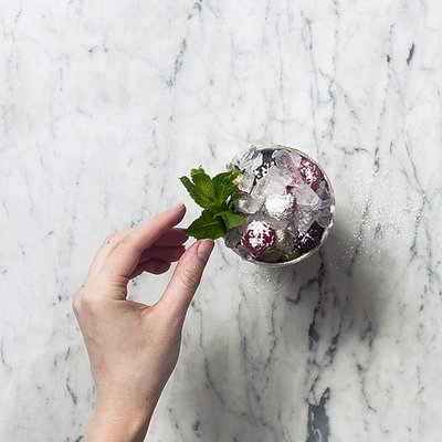 Eine Hand berührt ein Glas mit Eis und Beeren, umgeben von frischen Minzblättern auf einem marmorierten Tisch.
