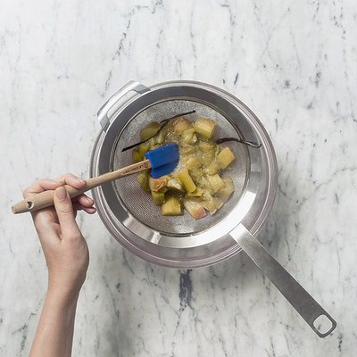 Eine Hand hält einen Holzlöffel und rührt in einem Sieb mit einer Mischung aus Rhabarber und Vanilleschoten.