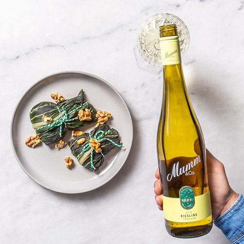 Ein Hand hält eine Flasche Mumm Riesling, während ein Teller mit gefüllten Blättern und Walnüssen im Hintergrund steht.