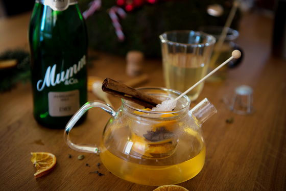 Auf dem Tisch liegt eine Teekanne mit einem aromatischen Getränk, umgeben von Zimt, getrockneten Orangenscheiben und Gläsern.