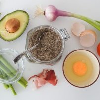 Auf dem Tisch sind verschiedene Lebensmittel angeordnet, darunter eine Avocado, Spargel, Zwiebeln und ein Glas mit Chiasamen.