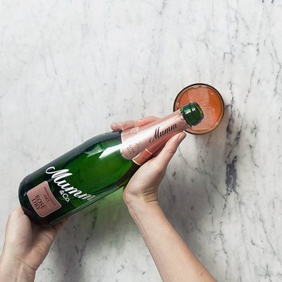 Eine Hand hält eine Flasche Mumm-Sekt über einem Glas mit einem rosafarbenen Getränk auf einer marmorierten Oberfläche.