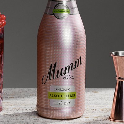 Eine Flasche alkoholfreier Rosé-Sekt neben einem dekorativen Glas voller Eis und frischen Beeren, auf einem hellen Tisch platziert.