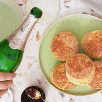 Eine Hand hält eine Flasche Sekt über einem Teller mit goldbraunen, fluffigen Brötchen, die appetitlich angerichtet sind.