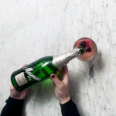 Eine Person hält eine Flasche Mumm-Sekt über einem Cocktailglas mit einer fruchtigen Mischung und Beeren.