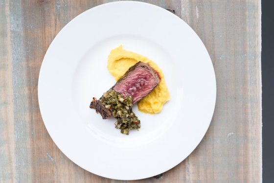 Ein saftiges Stück Rindfleisch liegt auf einem weißen Teller, garniert mit einer würzigen Kräutersauce und cremigem Polenta.