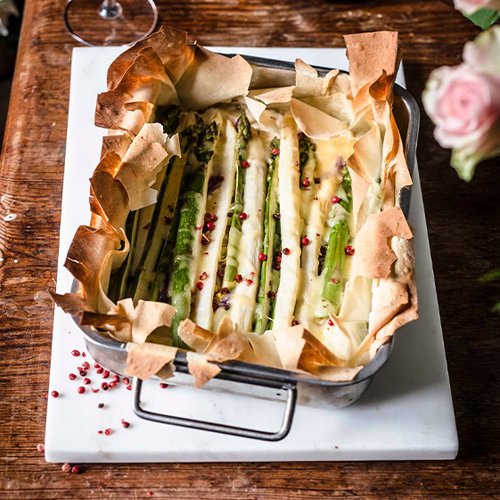 Ein rechteckiger Auflauf mit grünem Spargel, umhüllt von knusprigem Teig und garniert mit rosa Pfefferkörnern.