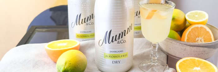 Drei Flaschen alkoholfreier Sekt stehen auf einem Tisch, umgeben von frischen Zitrusfrüchten und einem Glas mit einem Cocktail.