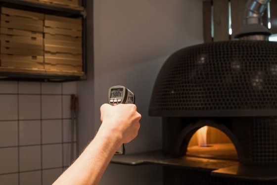 Eine Hand hält ein Infrarot-Thermometer, während ein Pizzaofen im Hintergrund sichtbar ist, der eine warme Glut ausstrahlt.