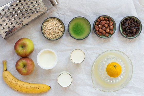 Eine Auswahl an frischen Zutaten liegt auf einem Tisch, darunter Äpfel, eine Banane, Haferflocken und Nüsse in Schalen.