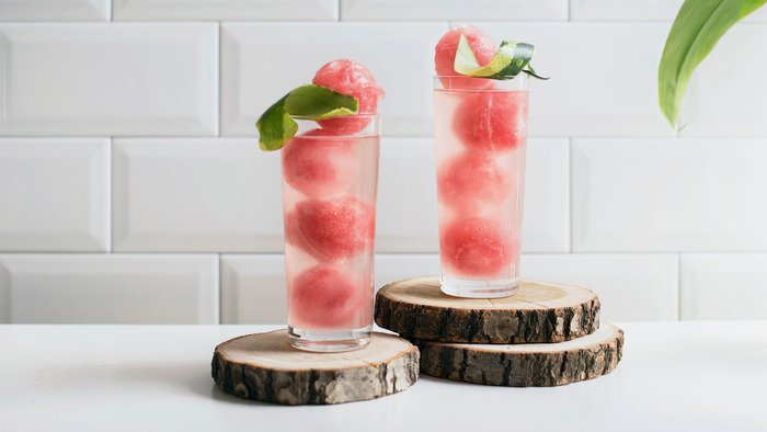 Zwei hohe Gläser mit Wassermelonen-Eis und frischen Blättern auf Holzscheiben, vor einer hellen Kachelnwand platziert.