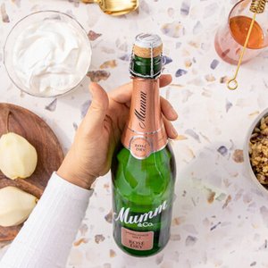 Die Flasche Mumm Rosé Dry steht im Mittelpunkt, während eine Hand sie greift, umgeben von frischen Birnen und anderen Leckereien.