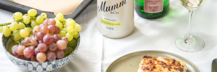 Ein elegantes Tischarrangement mit einer Schüssel Trauben, alkoholfreiem Sekt und einem köstlichen Stück Zwiebelkuchen auf einem Teller.