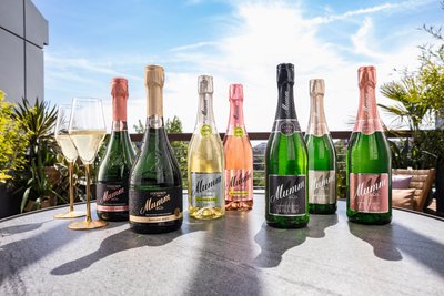 Sieben Flaschen Mumm Sekt in verschiedenen Desgins und Farben stehen auf einem grauen Untergrund, im Hintergrund ist ein sonniger Himmel zu sehen