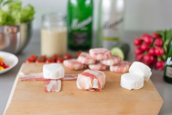 Eine appetitliche Anordnung von Speck umwickelten Käseportionen auf einem Schneidebrett, mit frischem Gemüse im Hintergrund.