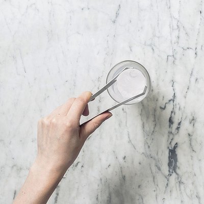 Eine Hand hält eine Zange, die einen Eiswürfel aus einem Glas auf einem marmorierten Tisch hebt.