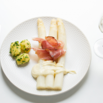Ein eleganter Teller mit weißen Spargeln, umgeben von Schinken, Kartoffeln und einer cremigen Sauce, serviert mit einem Glas Sekt.