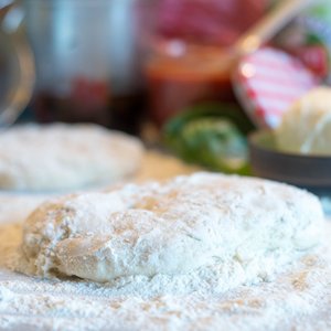 Auf einer bemehlten Arbeitsfläche liegen zwei Teigstücke, umgeben von verschiedenen Zutaten und Küchenutensilien.