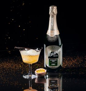 Ein eleganter Cocktail in einem Kristallglas, begleitet von einer Flasche Mumm Sekt und einer Passionsfrucht auf dem Tisch.
