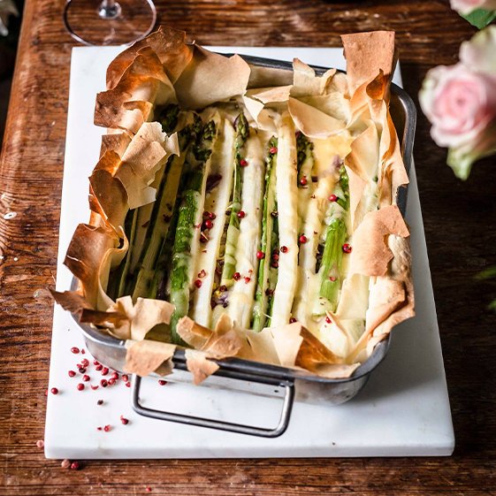 Ein rechteckiger Auflauf mit grünem Spargel, umhüllt von knusprigem Teig und garniert mit rosa Pfefferkörnern.