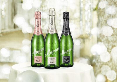 Auf einem weißen Tisch sind drei Flaschen Mumm Sekt platziert, jede mit einem anderen Etikett und funkelndem Hintergrund.