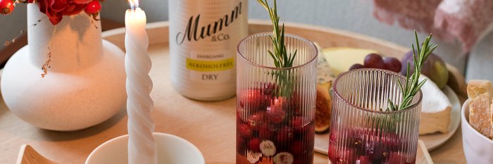 Auf einem Holztablett stehen zwei Gläser mit roten Früchten, eine elegante Kerze und eine Vase mit frischen Blumen in voller Blüte.