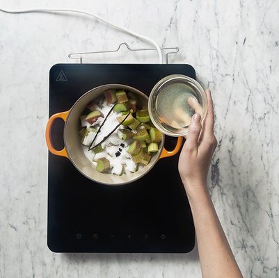 Eine Hand hält ein Glas mit Flüssigkeit über einen Topf mit Rhabarberstücken und Zucker auf einem Induktionskochfeld.