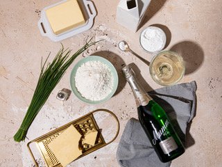 Eine Anordnung von Kochzutaten, darunter Mehl, Butter, Schnittlauch und ein Glas Sekt auf einer neutralen Oberfläche.