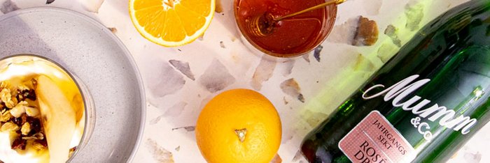 Ein elegantes Dessert mit Joghurt, Nüssen und Honig, umgeben von einer Orange und einer Flasche Sekt auf einem marmorierten Tisch.